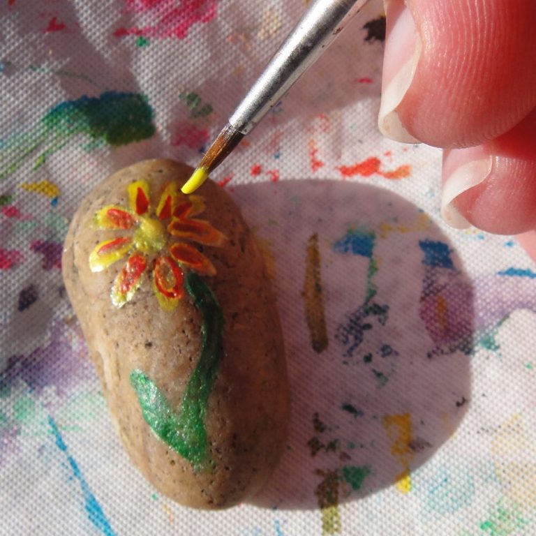 Pass the Pebble Photo of a stone being painted with the image of a flower, there is a fine paintbrush in the image loaded with yellow paint for the petals