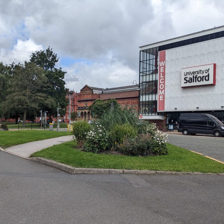 Re-Imaging the Green Door to Campus Photo of a grassy area in front of a large building with a 'Salford University' sign, there are plants and a path through the grass