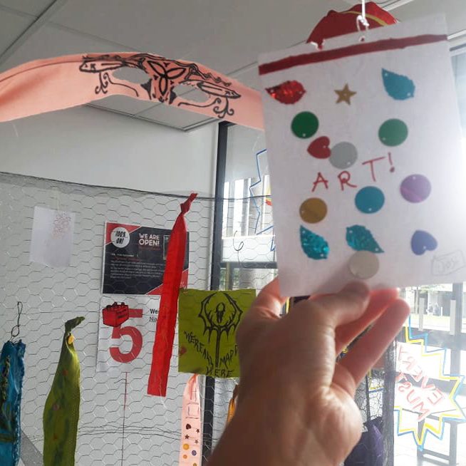 We Are Open Photo of a structure hung with coloured strips of fabric, a hand holding a piece of paper saying 'art!' childishly decorated with sequins