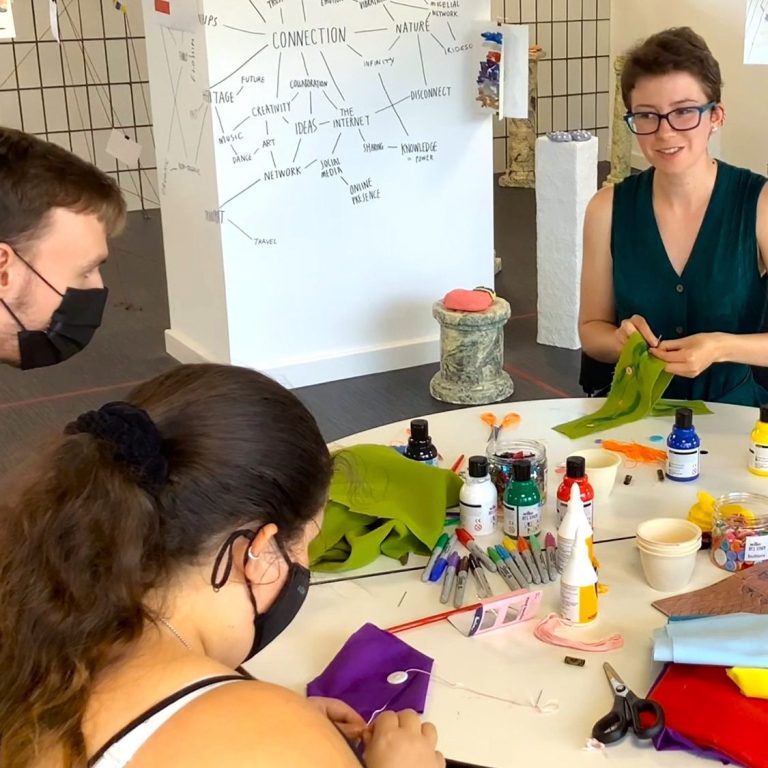 We Are Open Photo of a structure hung with coloured strips of fabric, a hand holding a piece of paper saying 'art!' childishly decorated with sequins